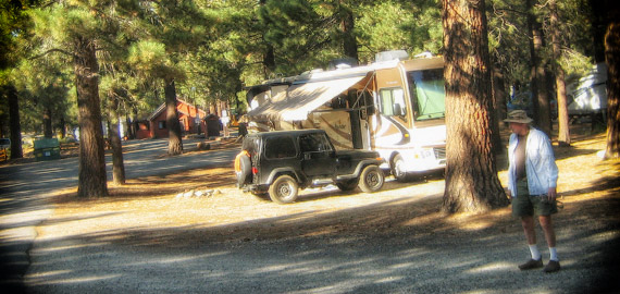 A motorhome and Jeep parked on a shaded site among tall pines at Mammoth Mountain RV Park, with a guest walking past in the late afternoon light.